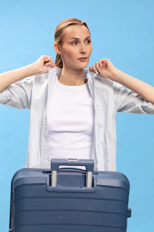Young woman with blonde hair stands behind blue suitcase adjusting shirt collar on blue background. Concept of travel preparation, packing, and casual clothing for trip or vacation.