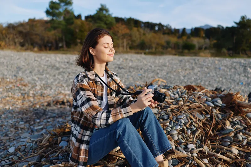 Camera loving woman sits on a rocky beach in a plaid shirt and jeans, enjoying a peaceful outdoor moment and capturing light, texture, and memories with her lens.