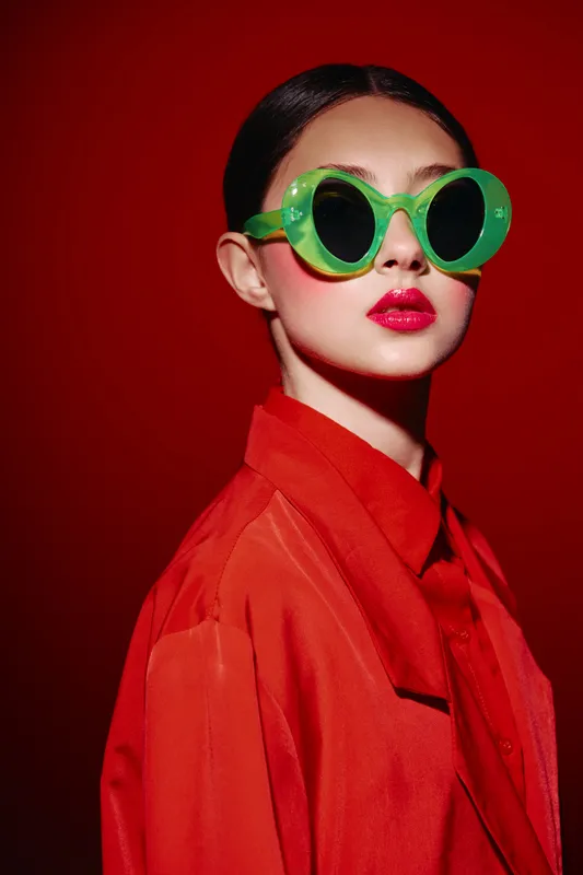 Young woman in oversized green sunglasses and vibrant red shirt poses confidently. Creative fashion portrait on red background with bright makeup and glossy lips. Concept of beauty and style.