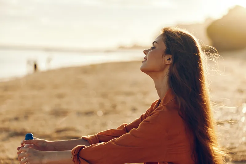 Joyful woman on a beach at sunset, relaxed pose, eyes closed, warm light, soft breeze, water bottle in hand, sandy shore and distant waves creating a serene scene