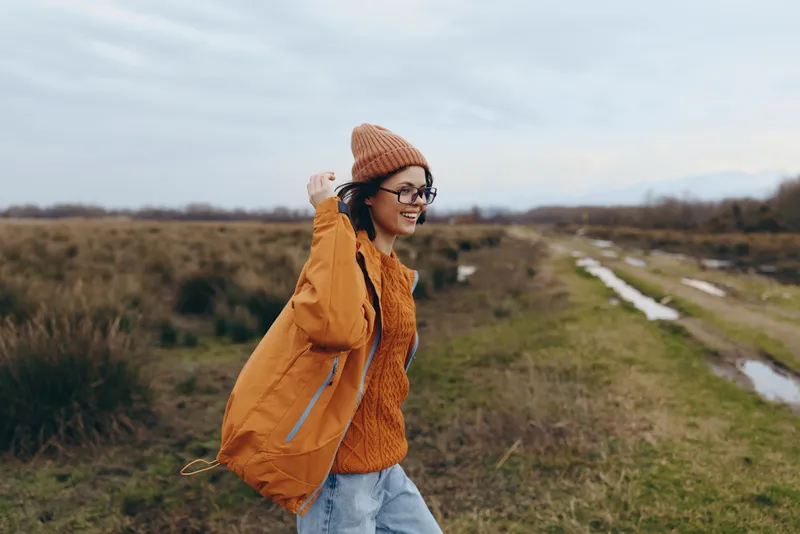 Woman hiking outdoors along a countryside path, wearing beanie, coat, and scarf. Autumn walking lifestyle on a rural landscape with grass, trees, and cloudy sky. Soft light in open fields.
