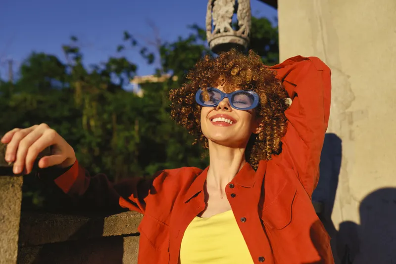 Smiling woman with curly hair wearing a red jacket and yellow top outdoors under bright sun. Relaxed lifestyle moment, warm light, carefree mood and confidence