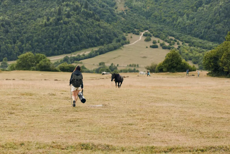 A lone hiker walks across a dry meadow toward a distant hillside as horses graze and distant figures appear, creating a tranquil rural scene with expansive skies ahead.