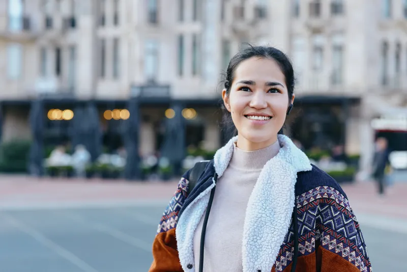 woman with warm smile wearing a cozy scarf and jacket, outdoor cityscape background, fashion forward casual look, confident demeanor, urban street scene, daytime