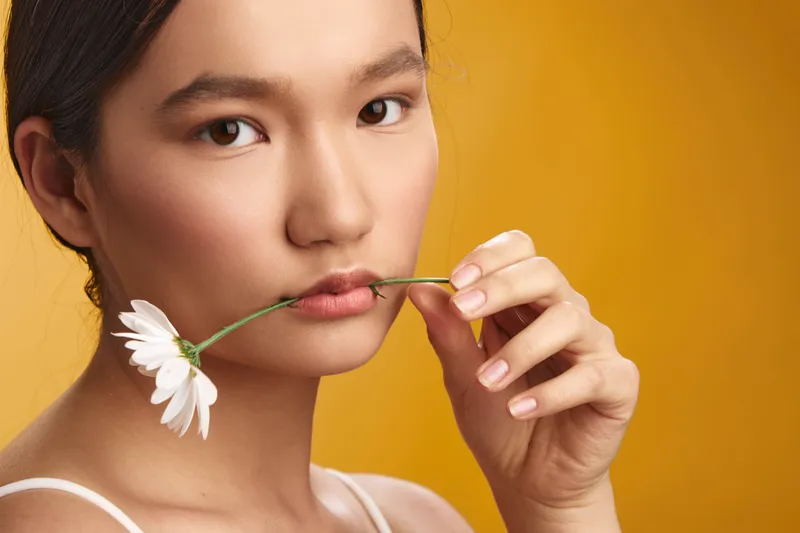 asian woman holds a flower stem near her lips in a soft studio setting. her calm gaze and gentle smile convey elegance, grace, and a serene beauty portrait.