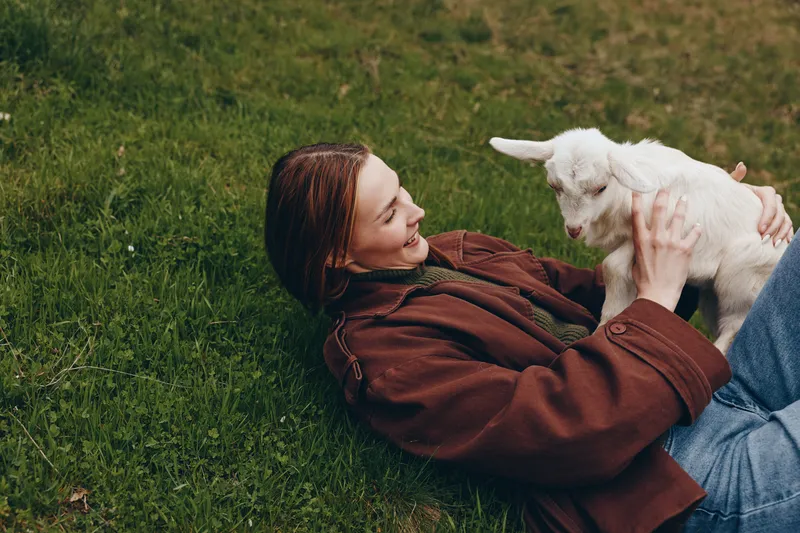 Young woman lying on lush grass in a brown jacket plays with a white goat kid, enjoying a joyful outdoor moment with gentle laughter and connection, in a bright natural setting.