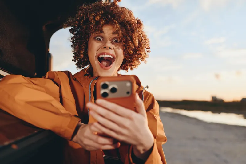 Smiling woman with curly hair taking a selfie with a smartphone, outdoors at the beach during sunset, lifestyle moment, orange jacket, laughter expression, soft evening light