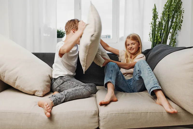 Two children having fun pillow fighting on beige sofa in bright living room, enjoying playful moment and carefree childhood indoors with casual clothes.