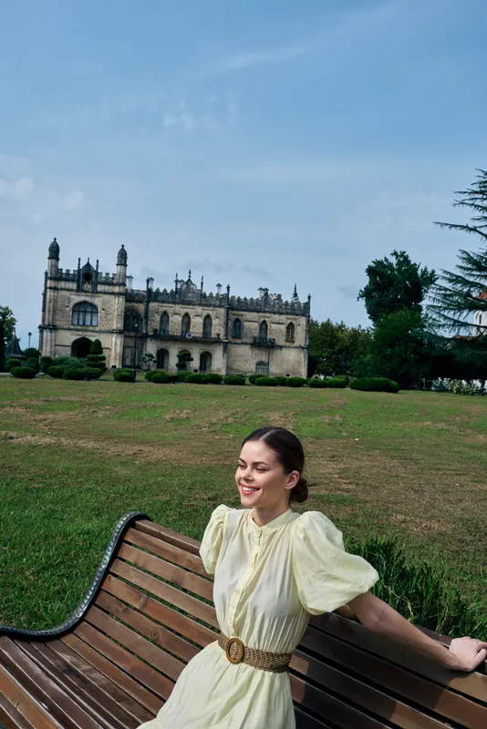 Woman in a pale dress sits on a bench in a green park with a historic castle in the background, smiling and enjoying a peaceful outdoor moment, on a sunny day, enjoying the scenery and calm ambiance.