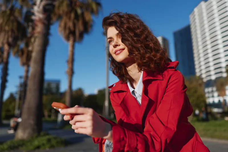 Young woman with curly hair in a red coat stands outdoors on a sunny city street, with palm trees and modern buildings in the background, smiling softly.