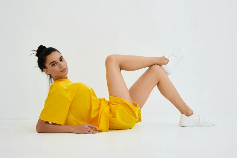 Young woman in yellow casual outfit reclining on white floor with relaxed expression, modern minimalistic background, natural light, sporty and fresh style