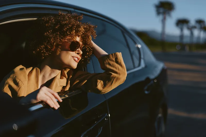 Woman leaning out of a vintage car window, sunlit breeze, carefree lifestyle moment, warm retro tones, casual fashion, outdoor road scene, natural colors, candid expression