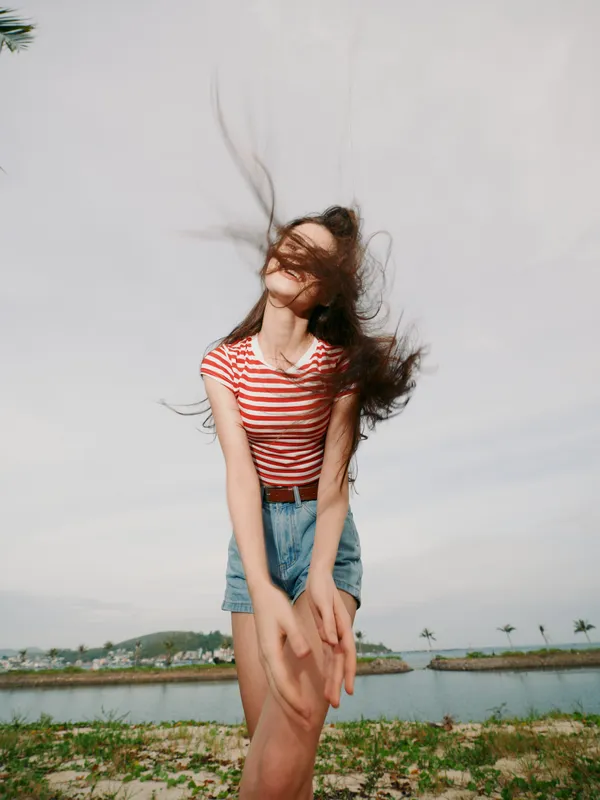 Young woman dances by a breezy shore, wearing a red striped tee and denim shorts, hair whirling in the wind, capturing a carefree, vibrant summer moment.
