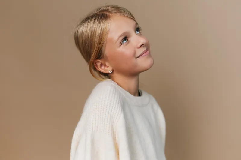 Portrait of a young woman in a soft white sweater, gazing upward with a gentle smile against a warm beige backdrop, conveying calmness and everyday elegance.