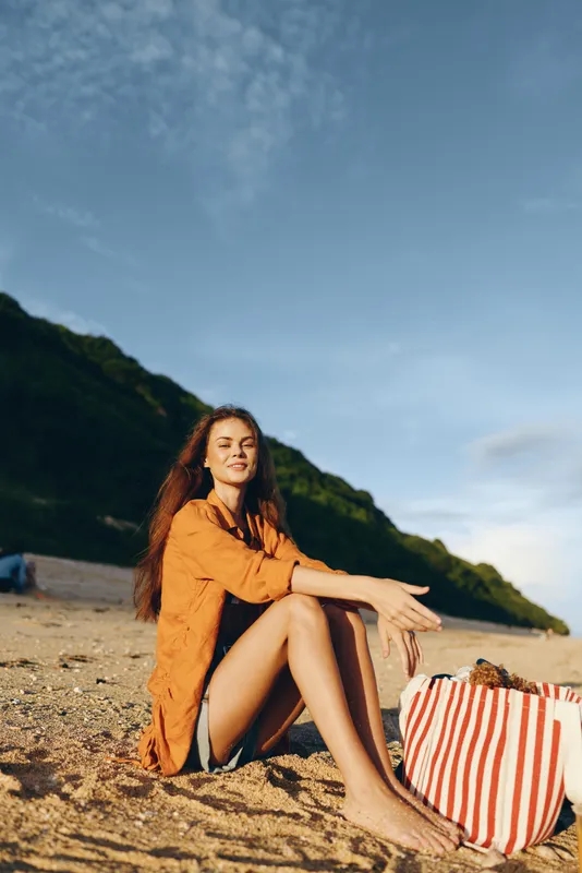 Young woman sits on a sunny beach in an orange shirt dress, smiling softly as the tide comes in and a striped tote sits beside her