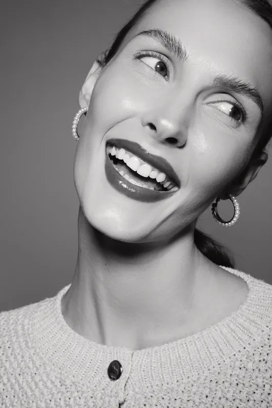 Smiling white caucasian woman with fair skin and glossy lips wears hoop earrings and knitted sweater. Concept of happiness, beauty, youth, and positive emotion in portrait studio.