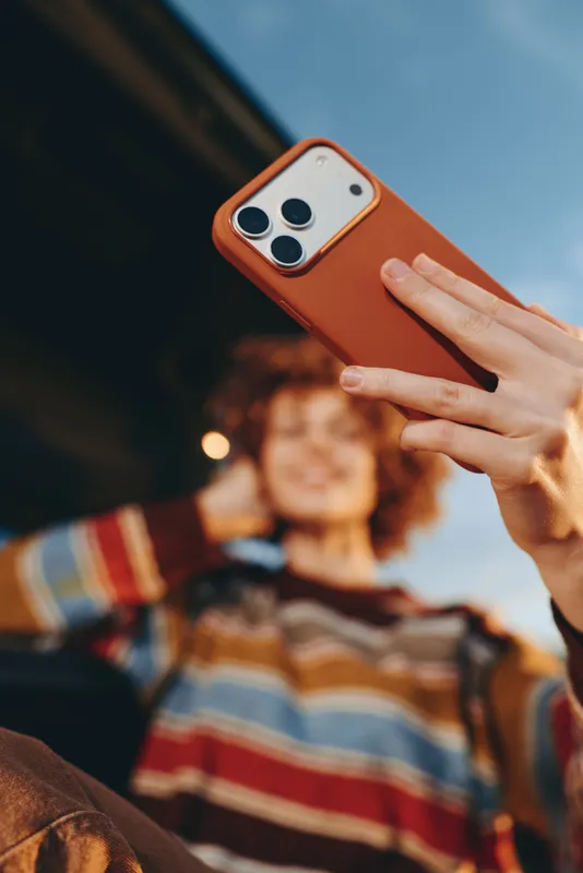 Woman lifestyle outdoors, hand holding orange smartphone for selfie, casual warm light, soft bokeh background with friends, communication and technology, closeup relaxed mood today