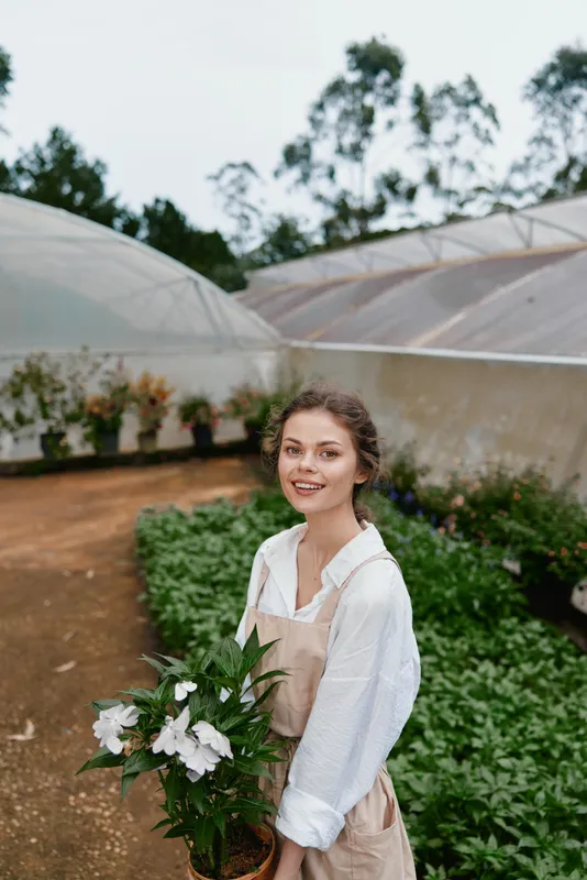 Young female gardener in an outdoor greenhouse holding flowers, wearing a white shirt and beige apron, smiling with greenery in the background