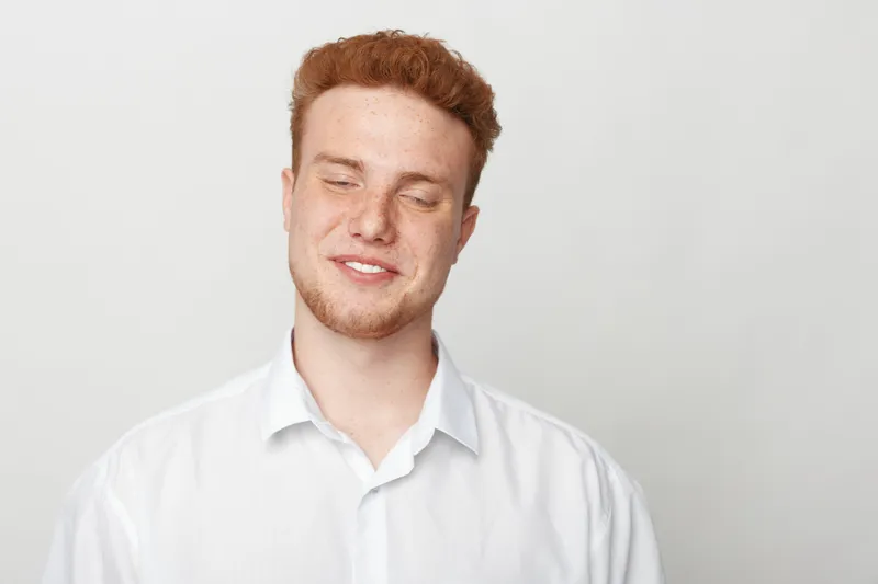 Young man with red hair and beard smiles gently with closed eyes wearing white shirt on light background. Concept of happiness, relaxation, and natural expression.
