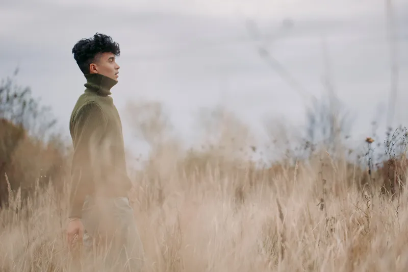 A genuine portrait of a man in a knit turtleneck standing in a windswept field, revealing authenticity and quiet confidence amid muted autumn tones.