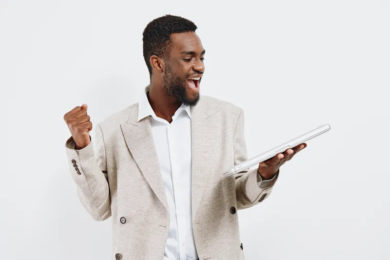 Confident smiling man of African descent in stylish beige blazer holding tablet device looking at screen isolated on plain white background studio portrait. People lifestyle concept