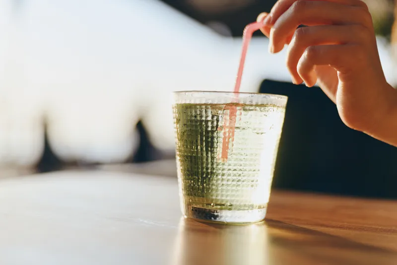 Lifestyle woman enjoying refreshing drink in modern boho-western denim street style, captured with warm film color tones and natural light on wooden surface.