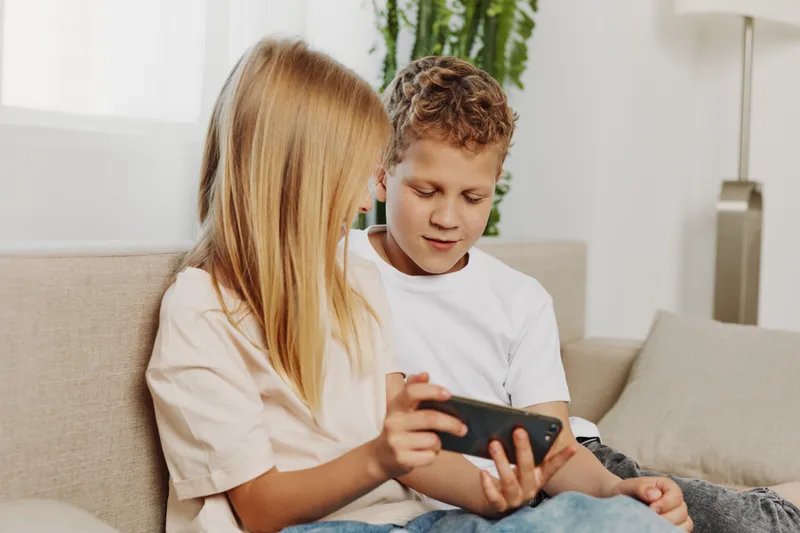Two children sit on couch playing with smartphone, focused and engaged in digital entertainment at home setting.