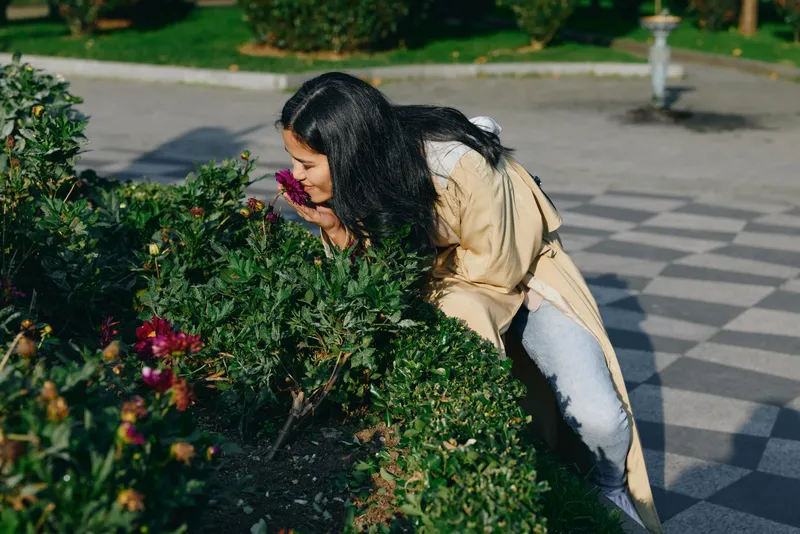 Woman bending to smell flowers in a garden, savoring the scent of blooms amid lush greenery, an outdoor scene focused on nature, wellness and calm, color