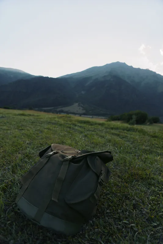 A green backpack sits on grassy terrain with mountains in the background under a clear sky. The scene represents outdoor adventure and nature exploration.
