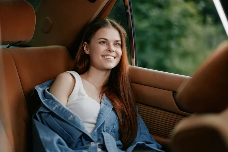Smiling girl in casual outfit inside a car, featuring long brown hair and a denim jacket, reflecting happiness and serenity in a natural outdoor setting.