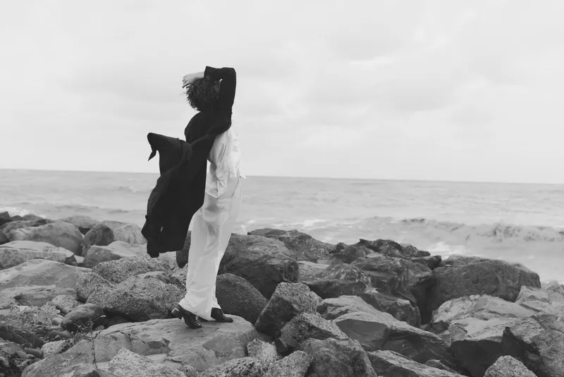 Couple embracing on a rocky shoreline by the sea, wind whipping garments, monochrome scene capturing love and resilience amid nature, outdoor romance, dramatic waves, rugged coast, timeless moment,