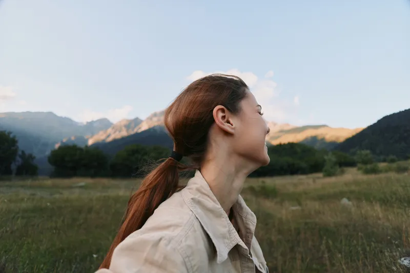 Young woman with long red hair tied in a ponytail enjoys a peaceful moment outdoors in a beige jacket with mountains and clear sky in the background.