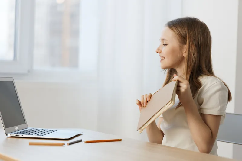 Joyful young woman smiling while holding a notebook near a laptop at a bright workspace, symbolizing creativity and motivation