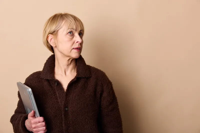 Mature elderly woman holding smartphone, surprised expression, reading message on beige studio background, indoor portrait, copy space for communication