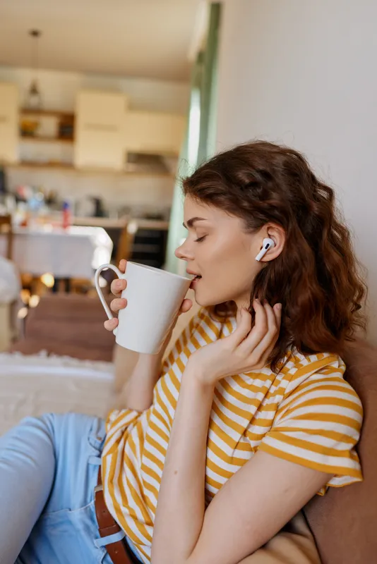 Relaxed woman at home in a striped tee sits against a wall, sipping from a mug, wearing wireless earbuds, enjoying a calm cozy lifestyle moment indoors in a sunlit living room