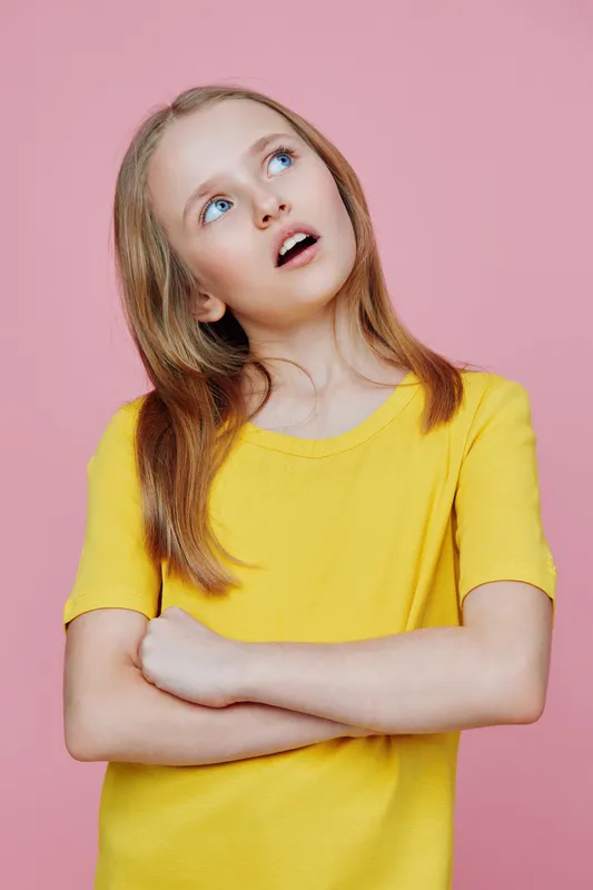 A thoughtful young girl with long hair wearing a bright yellow shirt, contemplating against a soft pink background, showcasing curiosity and innocence in her expression
