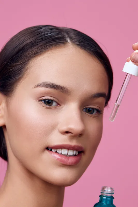 Young woman applying facial serum with dropper, showcasing healthy skin and natural beauty against a vibrant pink background