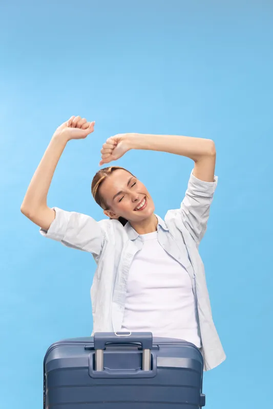 Happy young woman in casual clothes stretches arms up with eyes closed behind suitcase on blue background. Concept for travel, vacation, relaxation, and freedom.
