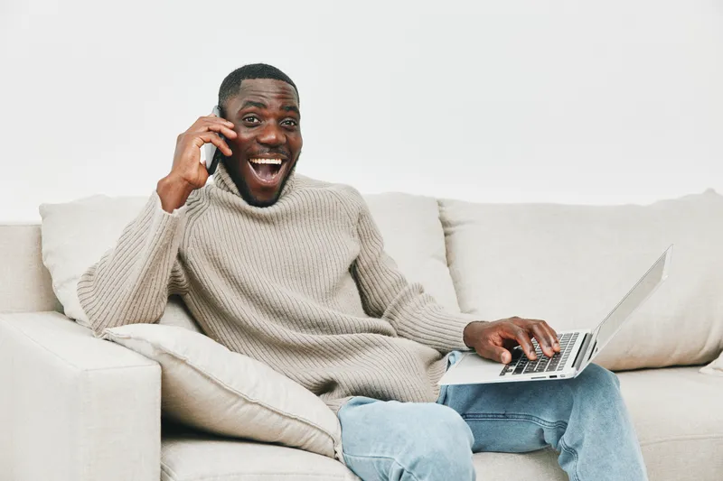 Young African man happily talking on the phone while using a laptop on a cozy sofa, expressing joy and excitement in a modern living room setting