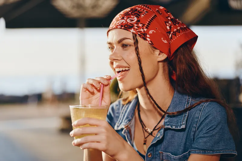 Lifestyle woman in modern boho-western denim street style enjoys a drink with a red bandana, smiling and relaxed, captured in warm film color tones.