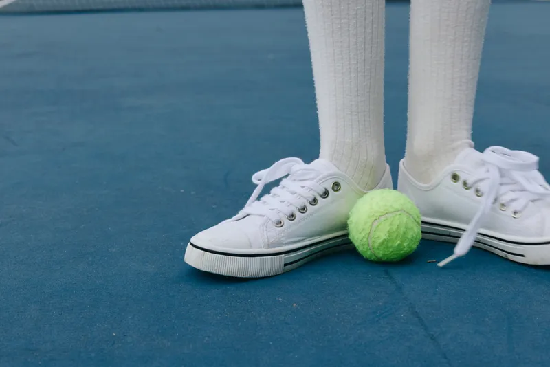 Young athlete in white sneakers standing on a tennis court, with a green tennis ball between the feet, representing sports and active lifestyle