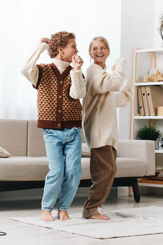 Two children dance indoors in cozy sweaters and jeans, smiling as they sway together, capturing carefree play and friendly rhythm in a bright, modern living room.