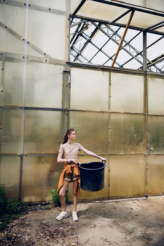 Woman greenhouse bucket gardening girl young carrying a large black bucket in a glasshouse, casual shorts and shirt, standing by translucent wall with sunlight, soil and horticulture tools.