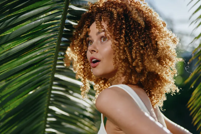Curly woman with surprised expression stands among tropical palm leaves in bright sunlight, wearing white sleeveless top during warm day outdoors.