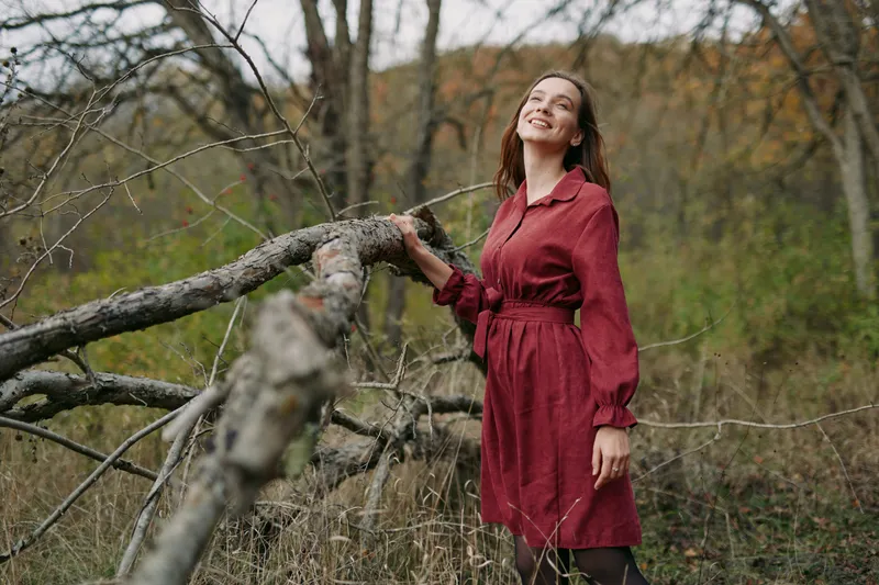 A calm woman in a burgundy dress stands beside a fallen branch in a forest, exuding authenticity and quiet confidence in natural light.