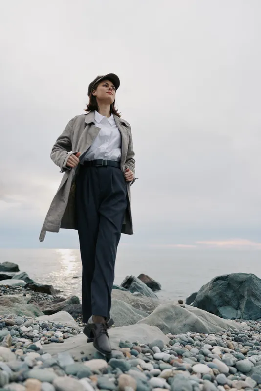 Young woman in trench coat and cap walks confidently on rocky beach under cloudy sky. Casual fashion and autumn style with sea and horizon in background. Outdoor lifestyle concept.