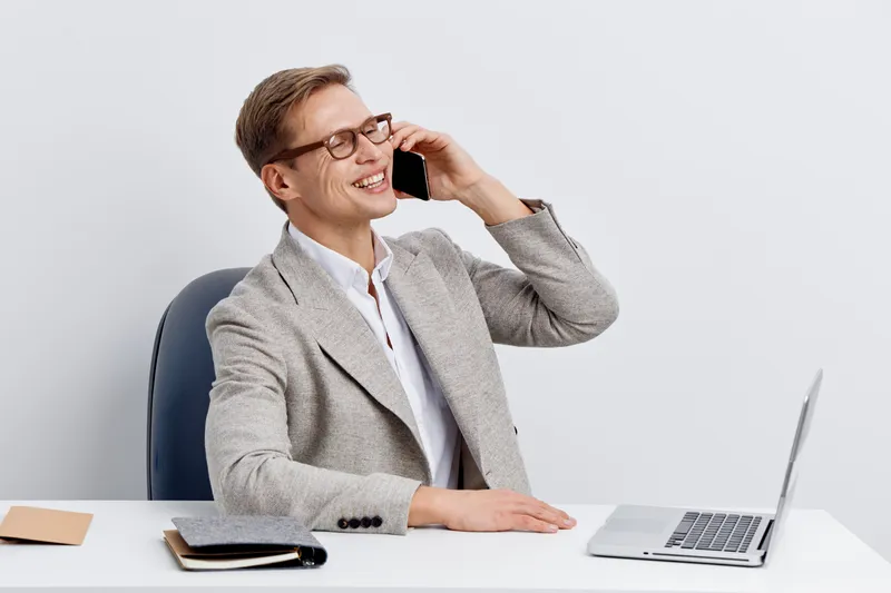 smiling young caucasian man chatting on smartphone with laptop at desk in light office casual jacket glasses positive emotion digital nomad concept