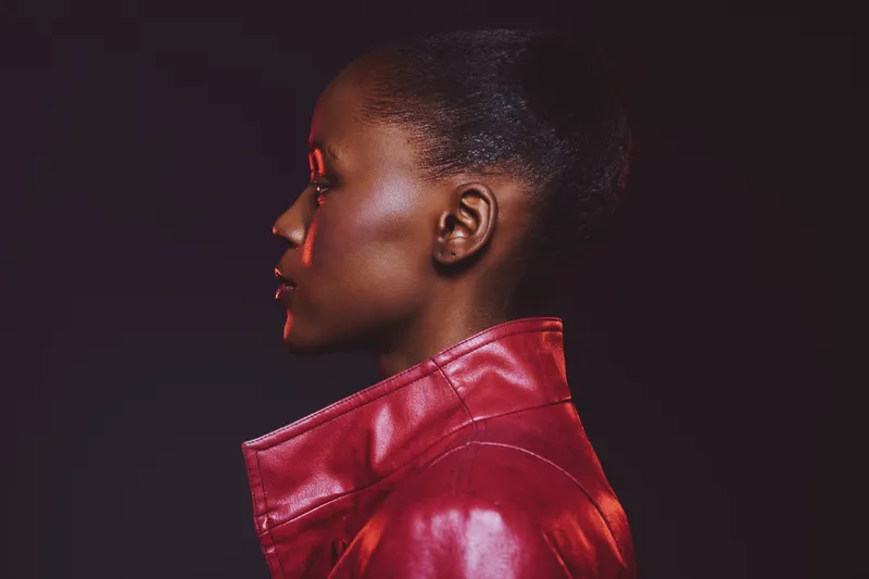 Profile of a dark skinned African woman wearing a red leather jacket. Studio portrait with dramatic lighting on black background. Concept for fashion, style, and beauty.
