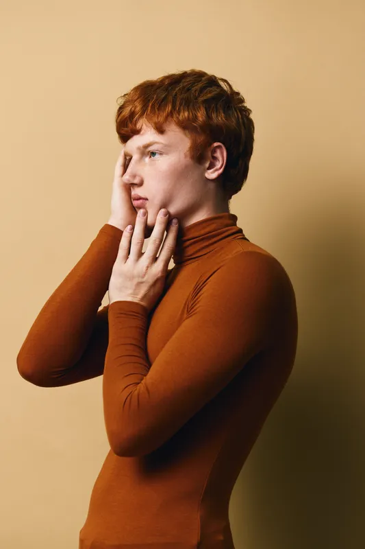 Young man with red hair wearing brown turtleneck poses thoughtfully with hand on face against beige background. Closeup portrait, emotional expression, modern fashion style.
