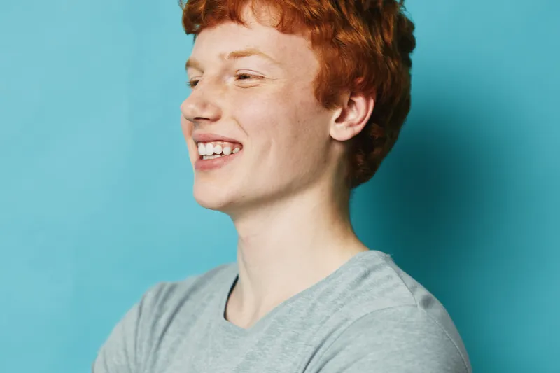 Young man with red hair smiling and looking sideways wearing casual gray t shirt on blue background. Portrait, happiness, youth and positive emotion concept.